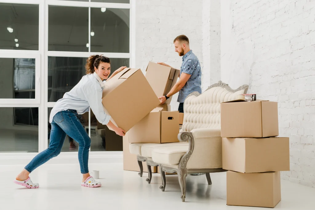 A couple carrying moving boxes past a sofa in a bright New York apartment