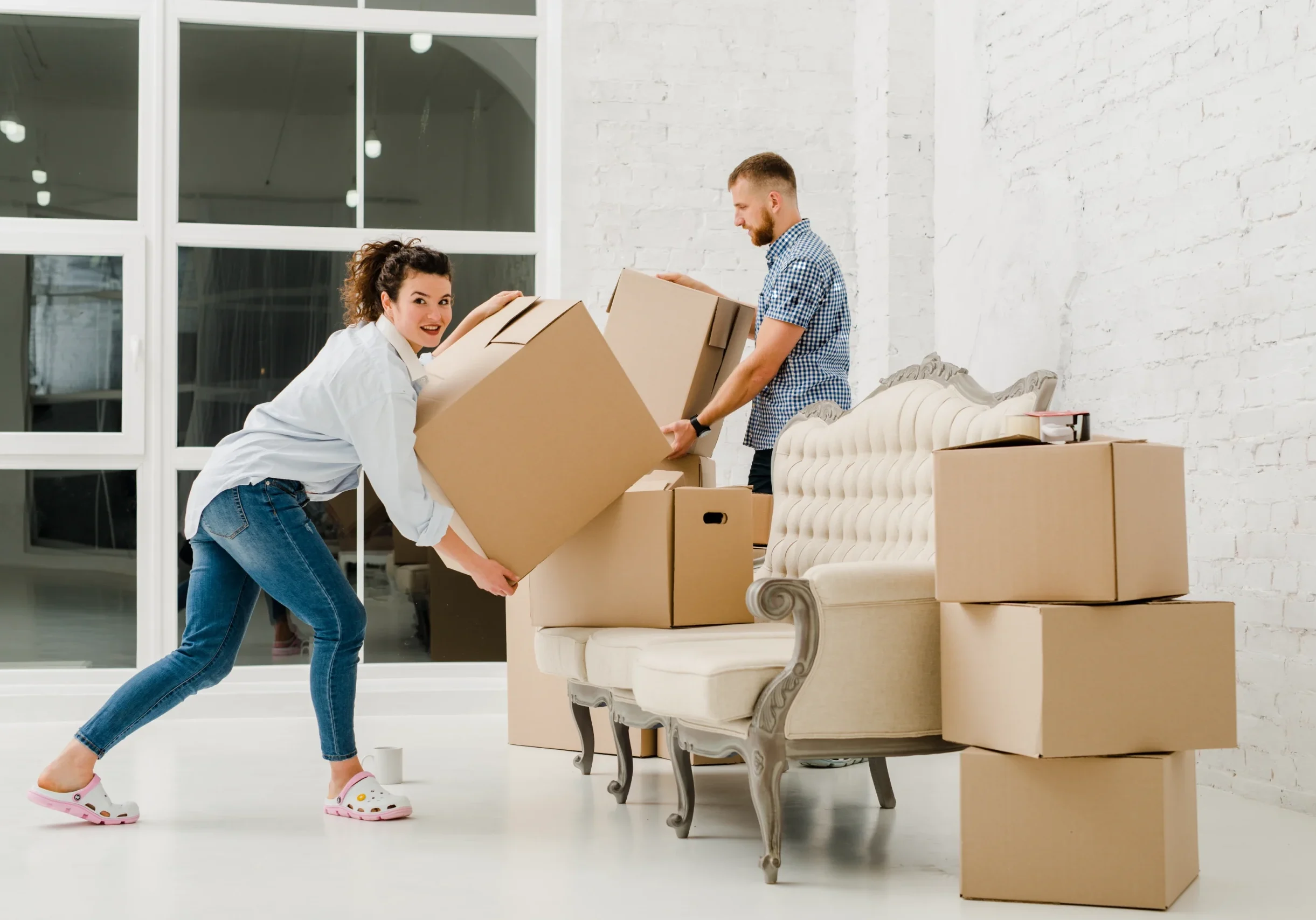 A couple carrying moving boxes past a sofa in a bright New York apartment